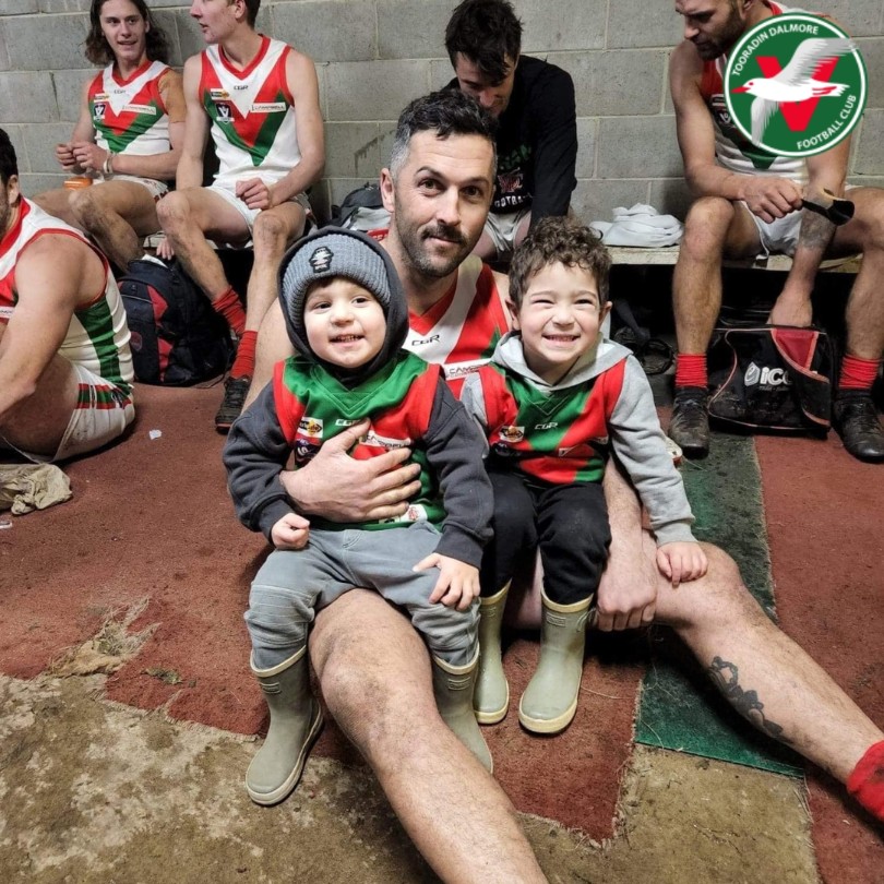 Football player sitting on the ground with two smiling kids on his knees wearing jumpers with the team colours of green and red.