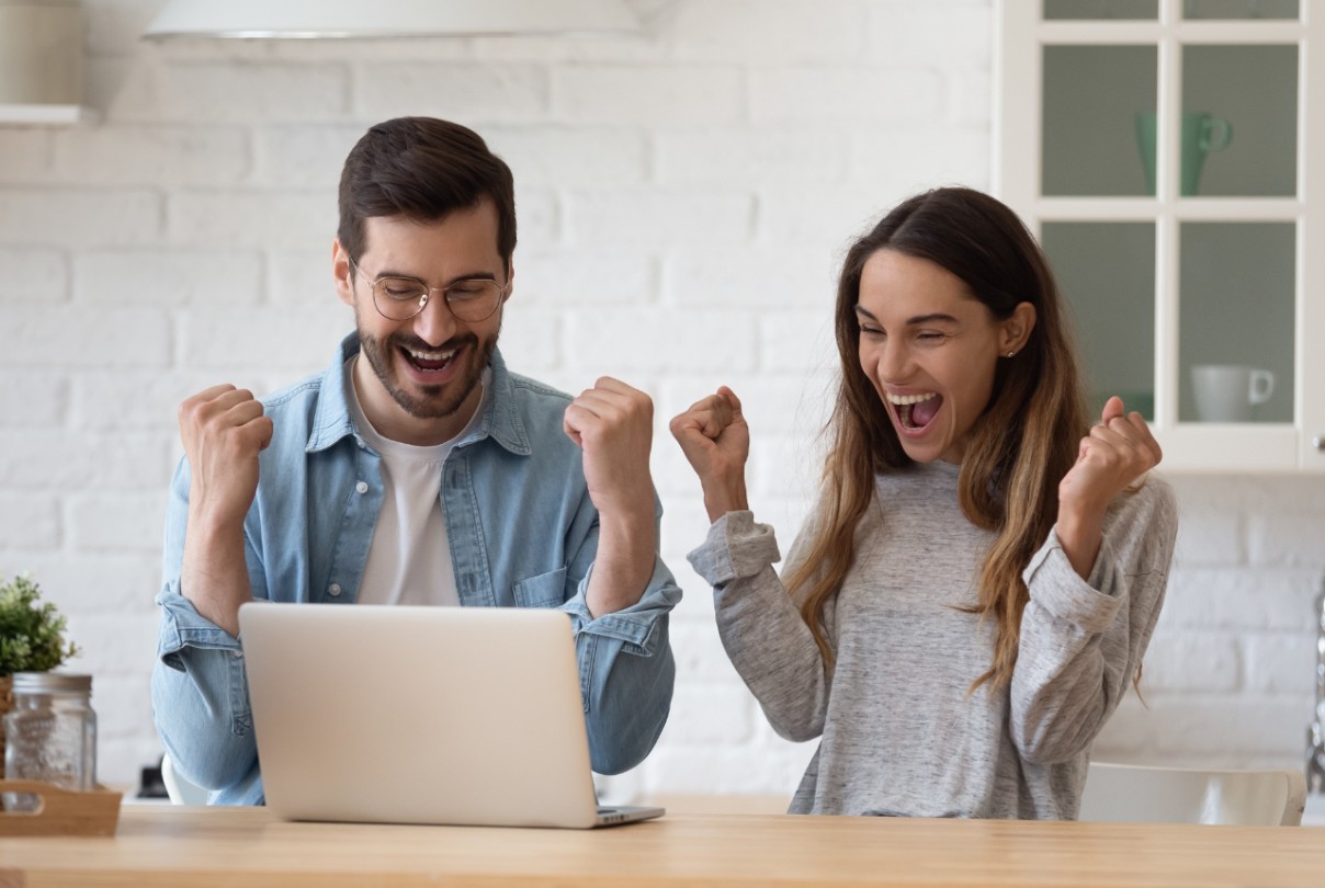 Excited young couple celebrating success, reading good news in email, looking at laptop screen, standing in modern kitchen at home, happy smiling woman and man showing yes gesture, online win