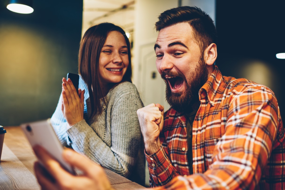 Happy marriage celebrating victory in internet lottery watching online broadcast on smartphone with winning results.Amazed hipsters wondered with win in online auction betting on website on phone