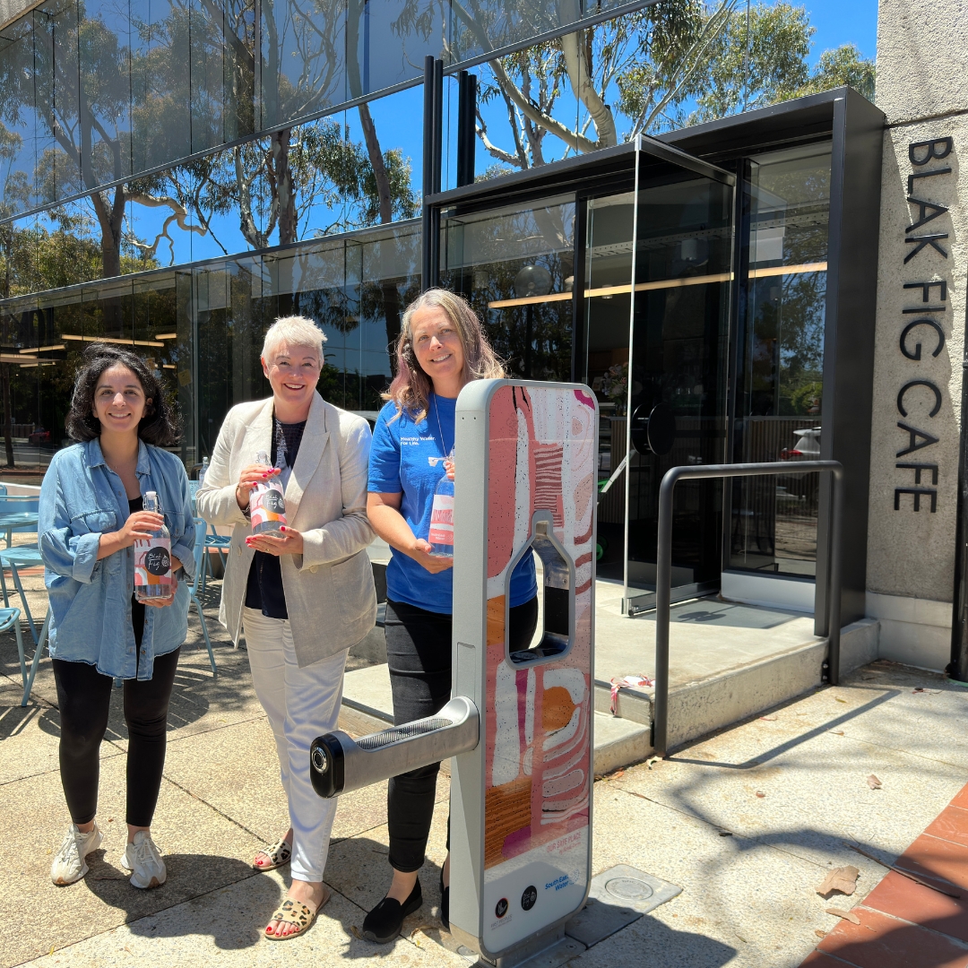 Three women smiling at the camera holding water bottles in front a water bottle refill station.