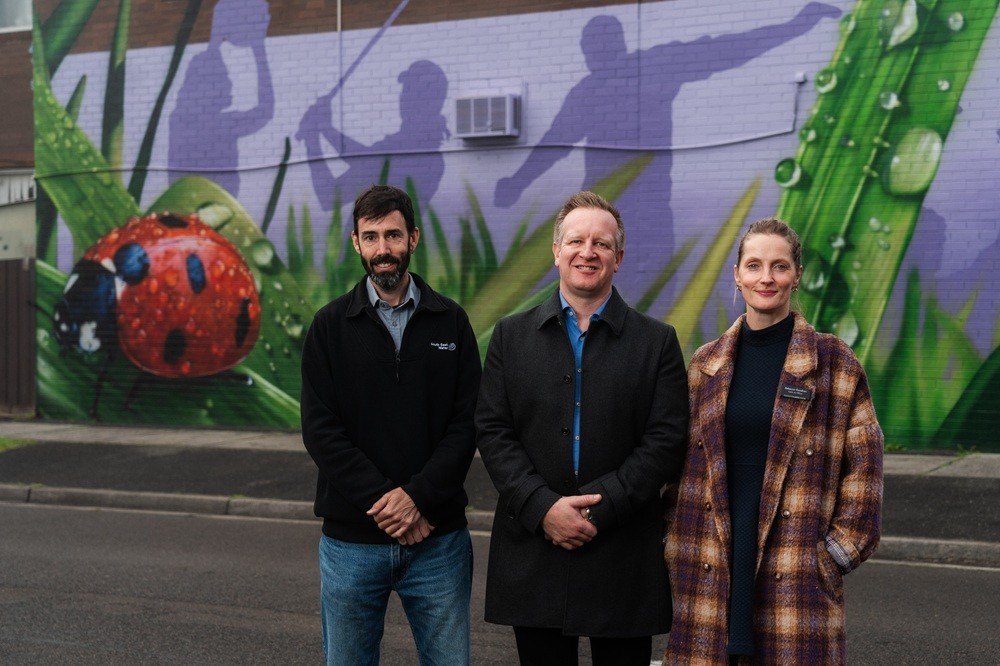 Three people standing outdoors in front of a colourful mural featuring large leaves and a ladybird. 
