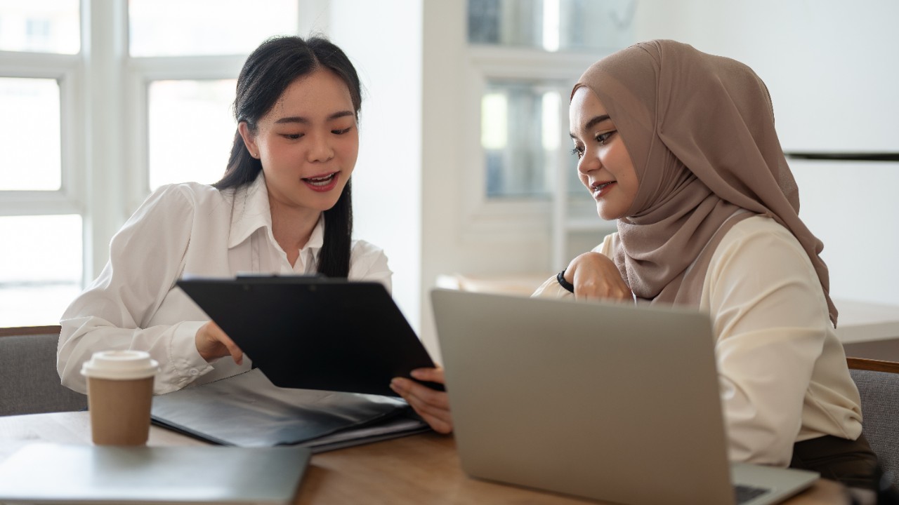 A beautiful Asian businesswoman is working with an Asian-Muslim female colleague in the meeting room, discussing work, brainstorming a project, asking questions, co-working.
